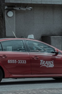 A red taxi with the branding 'Transcab' and a phone number is seen driving along a street. The vehicle is in motion, with a driver visible inside. The surroundings include a textured, gray stone building facade with some surveillance cameras mounted above.