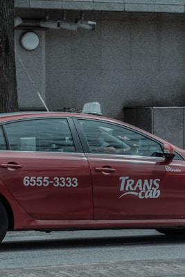 A red taxi with the branding 'Transcab' and a phone number is seen driving along a street. The vehicle is in motion, with a driver visible inside. The surroundings include a textured, gray stone building facade with some surveillance cameras mounted above.