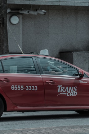 A red taxi with the branding 'Transcab' and a phone number is seen driving along a street. The vehicle is in motion, with a driver visible inside. The surroundings include a textured, gray stone building facade with some surveillance cameras mounted above.