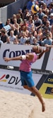 A beach volleyball player in mid-air during a jump serve, surrounded by a large, attentive crowd seated in the stands. The player is wearing a sleeveless pink shirt, blue shorts, and sunglasses. The volleyball is visible above the player's head, suggesting a moment of intense action. The audience is diverse, with many wearing sunglasses and summer attire, indicating a sunny day.