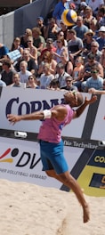 A beach volleyball player in mid-air during a jump serve, surrounded by a large, attentive crowd seated in the stands. The player is wearing a sleeveless pink shirt, blue shorts, and sunglasses. The volleyball is visible above the player's head, suggesting a moment of intense action. The audience is diverse, with many wearing sunglasses and summer attire, indicating a sunny day.