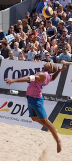 A beach volleyball player in mid-air during a jump serve, surrounded by a large, attentive crowd seated in the stands. The player is wearing a sleeveless pink shirt, blue shorts, and sunglasses. The volleyball is visible above the player's head, suggesting a moment of intense action. The audience is diverse, with many wearing sunglasses and summer attire, indicating a sunny day.