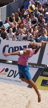 A beach volleyball player in mid-air during a jump serve, surrounded by a large, attentive crowd seated in the stands. The player is wearing a sleeveless pink shirt, blue shorts, and sunglasses. The volleyball is visible above the player's head, suggesting a moment of intense action. The audience is diverse, with many wearing sunglasses and summer attire, indicating a sunny day.