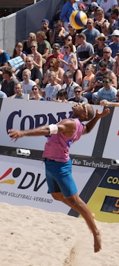 A beach volleyball player in mid-air during a jump serve, surrounded by a large, attentive crowd seated in the stands. The player is wearing a sleeveless pink shirt, blue shorts, and sunglasses. The volleyball is visible above the player's head, suggesting a moment of intense action. The audience is diverse, with many wearing sunglasses and summer attire, indicating a sunny day.