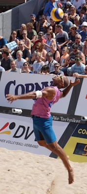 A beach volleyball player in mid-air during a jump serve, surrounded by a large, attentive crowd seated in the stands. The player is wearing a sleeveless pink shirt, blue shorts, and sunglasses. The volleyball is visible above the player's head, suggesting a moment of intense action. The audience is diverse, with many wearing sunglasses and summer attire, indicating a sunny day.