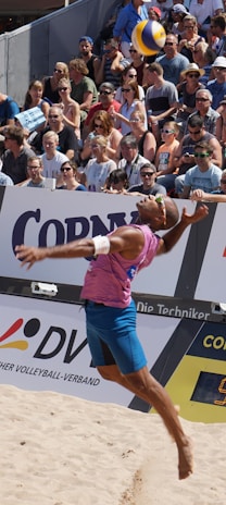A beach volleyball player in mid-air during a jump serve, surrounded by a large, attentive crowd seated in the stands. The player is wearing a sleeveless pink shirt, blue shorts, and sunglasses. The volleyball is visible above the player's head, suggesting a moment of intense action. The audience is diverse, with many wearing sunglasses and summer attire, indicating a sunny day.