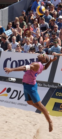 A beach volleyball player in mid-air during a jump serve, surrounded by a large, attentive crowd seated in the stands. The player is wearing a sleeveless pink shirt, blue shorts, and sunglasses. The volleyball is visible above the player