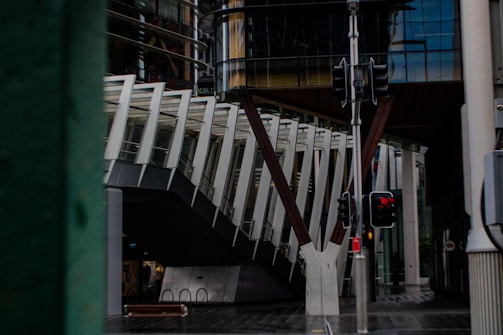 An urban street scene features a modern architectural structure with white, angled panels and large, brown triangular supports. A set of traffic lights stands in the foreground, and several buildings with reflective glass surfaces are visible in the background.