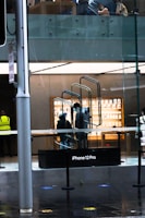 A modern store interior is visible through a glass facade, showcasing a display for the iPhone 12 Pro. Two people wearing masks are present inside, near a staircase. Outside, a person wearing a security vest stands by the entrance.