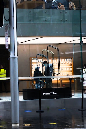 A modern store interior is visible through a glass facade, showcasing a display for the iPhone 12 Pro. Two people wearing masks are present inside, near a staircase. Outside, a person wearing a security vest stands by the entrance.