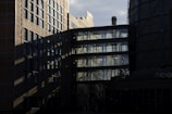A complex architectural structure with a mix of brick and glass elements is featured, casting intricate shadows. The reflection of the sky and structural elements can be seen in the glass windows, and a cylindrical tower with a chimney is partially visible.