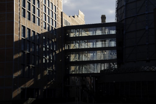 A complex architectural structure with a mix of brick and glass elements is featured, casting intricate shadows. The reflection of the sky and structural elements can be seen in the glass windows, and a cylindrical tower with a chimney is partially visible.