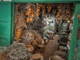An interior space filled with a variety of dried herbs and gourds hanging from the ceiling. Shelves are stocked with jars and bottles, possibly containing spices, teas, or herbal mixtures. The ground is covered with what appears to be mulch or dried material. The lighting is warm and inviting, casting an earthy and natural ambiance.
