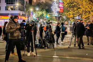 A group of people, mostly dressed in warm clothing, are gathered on a busy city street at night. Several of them are operating large video cameras mounted on tripods, suggesting a press or news event. The background is illuminated with city lights, including decorated trees and street signs. Some individuals are checking their phones while others are adjusting their equipment.