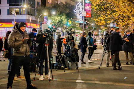 A group of people, mostly dressed in warm clothing, are gathered on a busy city street at night. Several of them are operating large video cameras mounted on tripods, suggesting a press or news event. The background is illuminated with city lights, including decorated trees and street signs. Some individuals are checking their phones while others are adjusting their equipment.