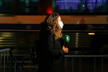 A person with curly hair is wearing a face mask and holding a green microphone with a logo, seemingly engaged in a television broadcast. They are standing near a barricade with blurred motion of a bus or train in the background, suggesting an urban setting at night or evening.
