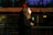 A person with curly hair is wearing a face mask and holding a green microphone with a logo, seemingly engaged in a television broadcast. They are standing near a barricade with blurred motion of a bus or train in the background, suggesting an urban setting at night or evening.