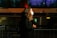A person with curly hair is wearing a face mask and holding a green microphone with a logo, seemingly engaged in a television broadcast. They are standing near a barricade with blurred motion of a bus or train in the background, suggesting an urban setting at night or evening.