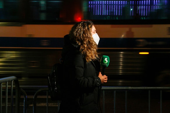 A person with curly hair is wearing a face mask and holding a green microphone with a logo, seemingly engaged in a television broadcast. They are standing near a barricade with blurred motion of a bus or train in the background, suggesting an urban setting at night or evening.