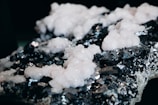 A close-up view of a mineral specimen with a rough, dark surface adorned with clusters of white, crystalline formations. The contrast between the light crystals and the dark, reflective background creates a striking visual effect.