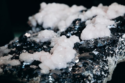 A close-up view of a mineral specimen with a rough, dark surface adorned with clusters of white, crystalline formations. The contrast between the light crystals and the dark, reflective background creates a striking visual effect.