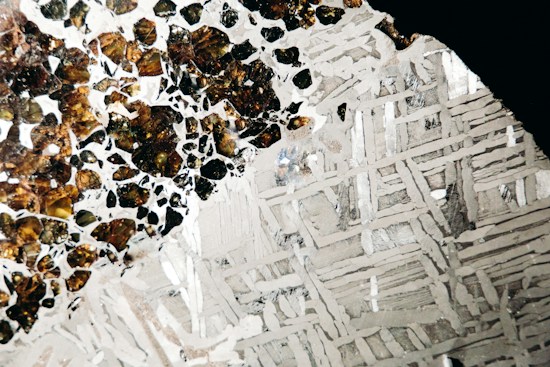A close-up image featuring a polished section of a meteorite highlighting its intricate metallic patterns. On one side, small, irregular, dark brown to black inclusions are embedded within a lighter matrix. The other half consists of silver-gray metallic crystals forming geometric patterns.