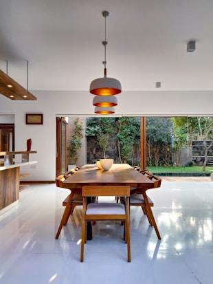 A modern dining area featuring a large wooden table surrounded by several matching chairs, all set on a glossy white floor. A contemporary pendant light hangs above the table, and a large window allows natural light to illuminate the space, providing a view of an outdoor garden with trees and greenery.