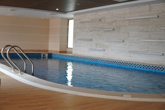 A serene indoor swimming pool with tiled walls and a wooden ceiling. The water in the pool is calm, reflecting the light from a nearby window. Stainless steel handrails are positioned at one end of the pool, and the floor surrounding the pool has a wood-like finish.