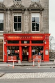 A vibrant red storefront named 'The Elephant House' with large windows and ornate architectural details. The sign indicates the location as the 'birthplace of Harry Potter.' The scene includes barriers on the road in front, and the interior appears welcoming with visible patrons inside.