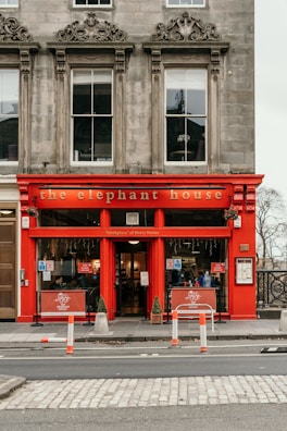 A vibrant red storefront named 'The Elephant House' with large windows and ornate architectural details. The sign indicates the location as the 'birthplace of Harry Potter.' The scene includes barriers on the road in front, and the interior appears welcoming with visible patrons inside.