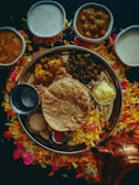 An array of traditional Indian dishes is arranged on a large round platter, surrounded by colorful flower petals. The platter includes flatbread, a dollop of butter, and various curries. Several small bowls containing creamy and spicy dishes are placed around the platter. A copper container is visible near the bottom right.
