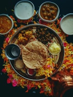 An array of traditional Indian dishes is arranged on a large round platter, surrounded by colorful flower petals. The platter includes flatbread, a dollop of butter, and various curries. Several small bowls containing creamy and spicy dishes are placed around the platter. A copper container is visible near the bottom right.