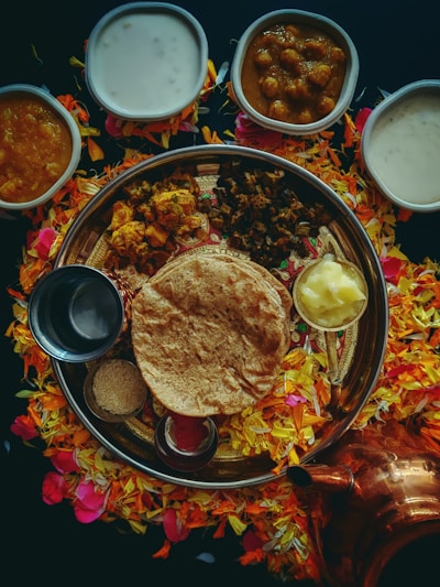An array of traditional Indian dishes is arranged on a large round platter, surrounded by colorful flower petals. The platter includes flatbread, a dollop of butter, and various curries. Several small bowls containing creamy and spicy dishes are placed around the platter. A copper container is visible near the bottom right.