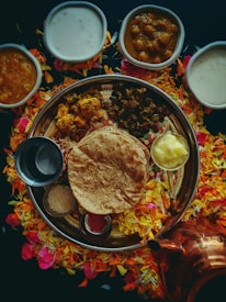 An array of traditional Indian dishes is arranged on a large round platter, surrounded by colorful flower petals. The platter includes flatbread, a dollop of butter, and various curries. Several small bowls containing creamy and spicy dishes are placed around the platter. A copper container is visible near the bottom right.