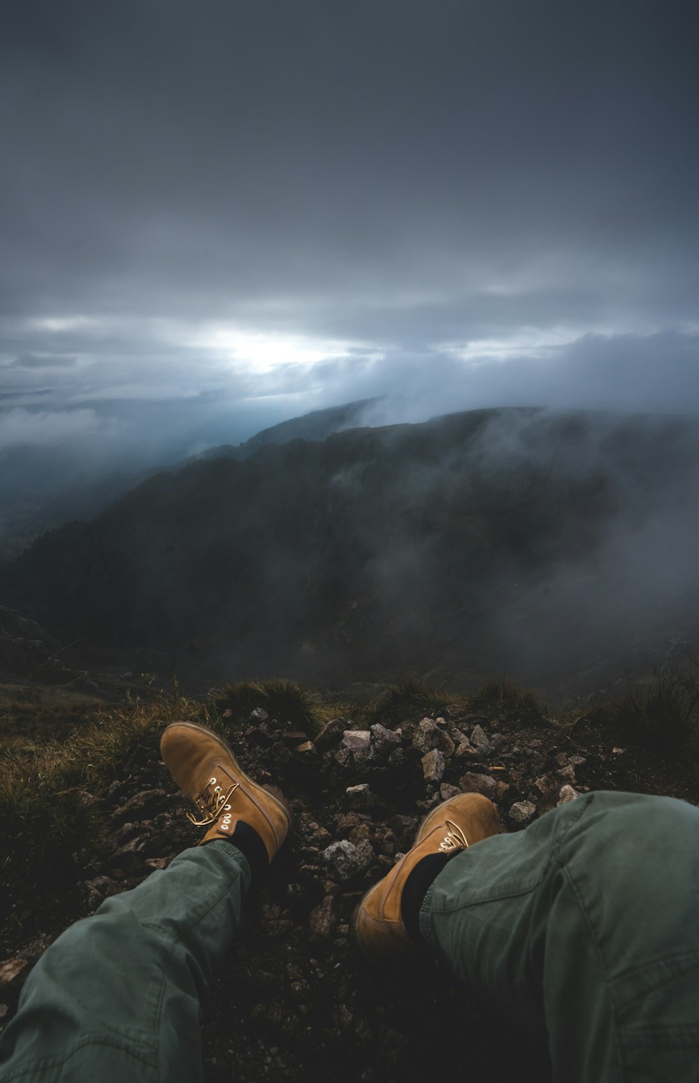 person in green pants and brown hiking shoes sitting on green grass field near green mountains