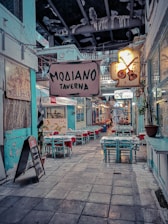 A narrow alleyway with a cozy atmosphere features several tables and chairs arranged outside a traditional-looking taverna. The sign above reads 'Modiano Taverna' and another illuminated sign displays musical instruments, suggesting a lively ambiance. The walls are painted in muted turquoise, paired with red tablecloths that add a splash of color. Hanging lights and potted plants contribute to the welcoming setting.