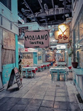 A narrow alleyway with a cozy atmosphere features several tables and chairs arranged outside a traditional-looking taverna. The sign above reads 'Modiano Taverna' and another illuminated sign displays musical instruments, suggesting a lively ambiance. The walls are painted in muted turquoise, paired with red tablecloths that add a splash of color. Hanging lights and potted plants contribute to the welcoming setting.