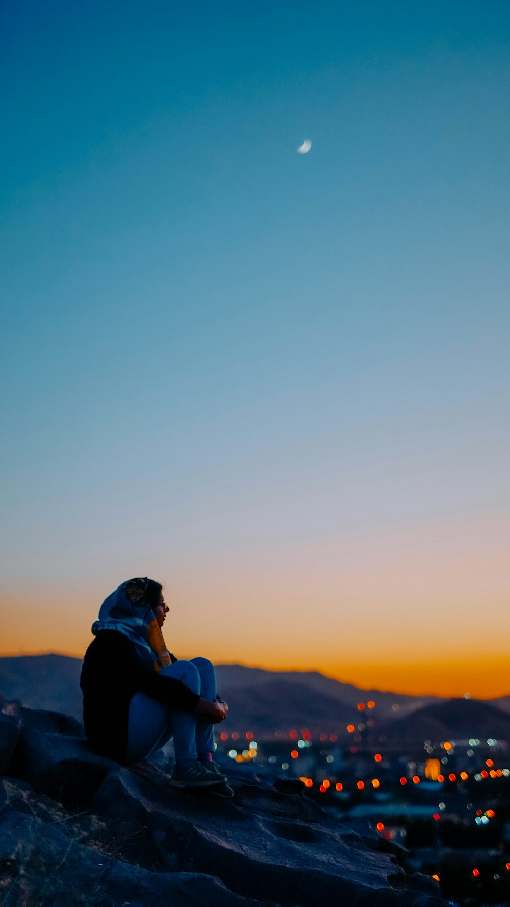 man in blue jacket and black cap sitting on rock during sunset