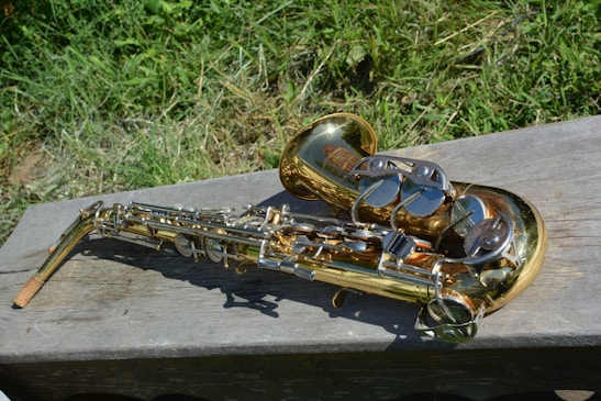 A golden saxophone placed on a wooden bench with grass in the background. The instrument's metallic surface reflects sunlight, and its intricate keys and structure are visible.