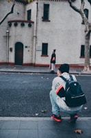 A person with a backpack squats on the roadside, seemingly taking a picture of another person who is walking on the other side of the street. The buildings in the background have a vintage architectural style with white walls and red trimming.