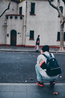 A person with a backpack squats on the roadside, seemingly taking a picture of another person who is walking on the other side of the street. The buildings in the background have a vintage architectural style with white walls and red trimming.