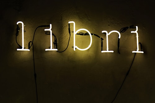 A neon sign displaying the word 'libri' in lowercase letters is mounted on a textured wall. The letters glow with a soft yellow light while black cables are visible connecting each letter.
