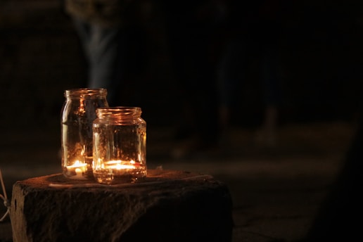 Two glass jars containing lit candles are placed on a stone surface. The background is dimly lit, creating a warm and serene ambiance.