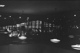 A dark, monochromatic interior scene featuring several ceiling lamps emitting circular light patterns. The background includes reflections on glass showcasing an urban environment with buildings and glimpses of ambient lighting.