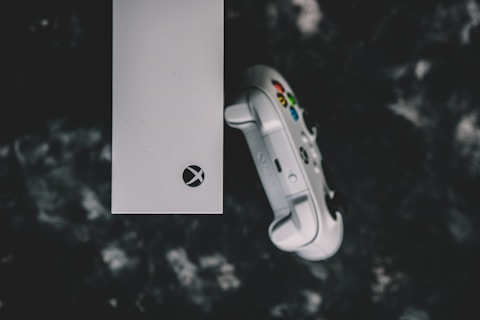 A top view of a white gaming console and a matching controller set against a dark, textured background. The console features a prominent logo, while the controller displays colorful buttons.