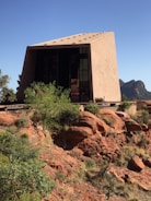 A modern architectural structure with a slanted roof stands on a rocky, red desert landscape. Surrounding the building are green shrubs and plants, with a clear blue sky in the background. The building features large glass windows reflecting the outdoors.