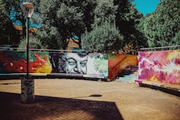 A vibrant street art mural is painted on a curved wall, featuring a large, black-and-white portrait of a man's face surrounded by abstract, colorful designs. The setting includes a cobblestone pavement with a lamppost casting a shadow. Trees with dense, dark green foliage provide a natural backdrop, and a set of stone stairs leads up behind the mural.