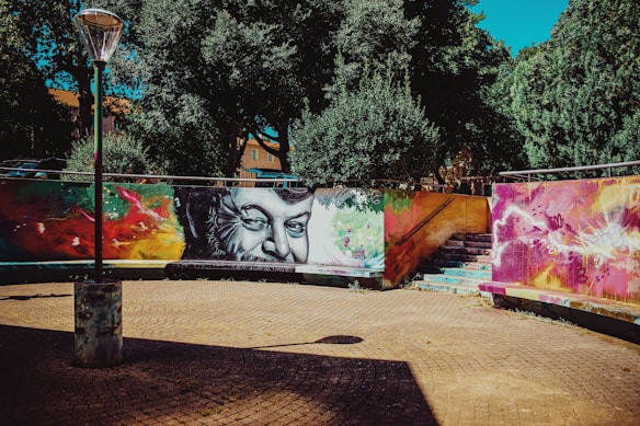 A vibrant street art mural is painted on a curved wall, featuring a large, black-and-white portrait of a man's face surrounded by abstract, colorful designs. The setting includes a cobblestone pavement with a lamppost casting a shadow. Trees with dense, dark green foliage provide a natural backdrop, and a set of stone stairs leads up behind the mural.