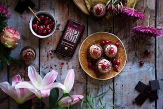 An arrangement of chocolate desserts garnished with cherries is placed on a decorated plate. Nearby, a bowl filled with cherries and sauce, an array of fresh flowers including lilies and roses, and a few pieces of dark chocolate are positioned on a rustic wooden surface. A smartphone displaying a podcast titled 'Bad Faith' is also present, contributing a modern touch to the food and floral setting.