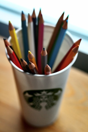 A collection of colorful wooden pencils is placed inside a paper cup. The cup features a distinct logo and sits on a wooden surface. The pencils vary in color, including red, yellow, blue, and orange, and are arranged with their sharpened tips pointing upward. A soft, diffused light brightens the scene, adding warmth to the overall atmosphere.
